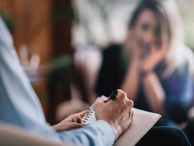 Therapist taking notes during a counseling session, client visible in the background blurred.