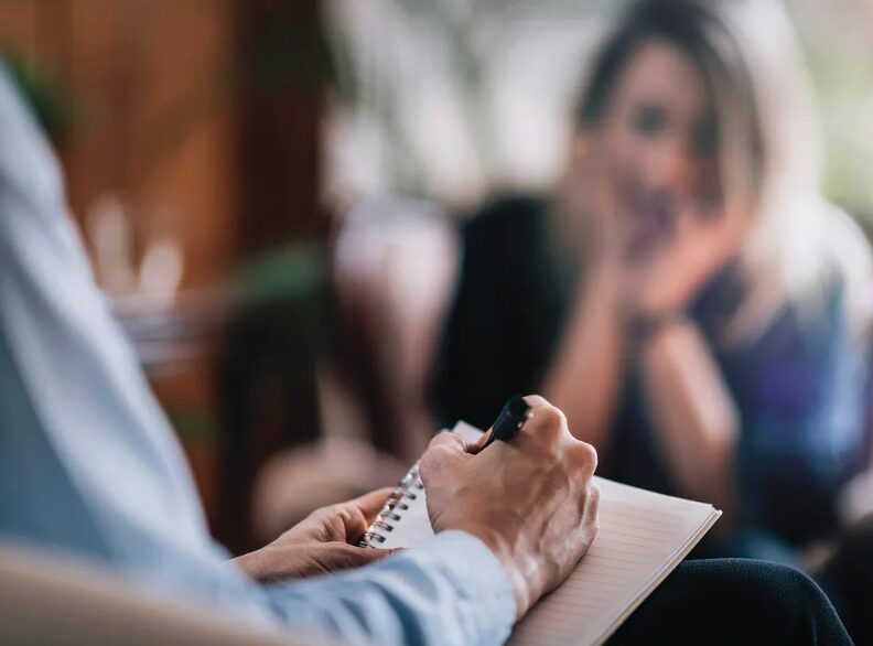 Therapist taking notes during a counseling session, client visible in the background blurred.