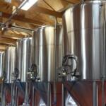 Row of large stainless steel fermentation tanks in a brewery, with pipes and wooden ceiling visible