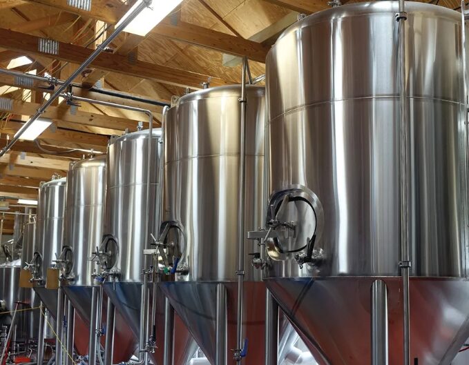 Row of large stainless steel fermentation tanks in a brewery, with pipes and wooden ceiling visible