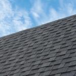 Diagonal view of a textured gray asphalt shingle roof against a blue sky with wispy clouds.