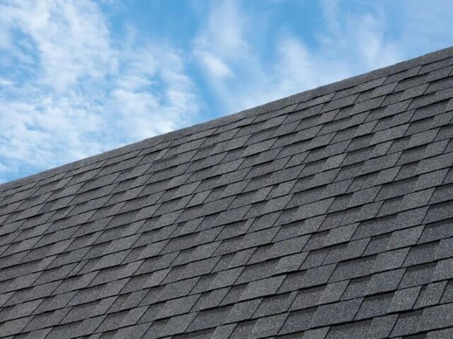 Diagonal view of a textured gray asphalt shingle roof against a blue sky with wispy clouds.