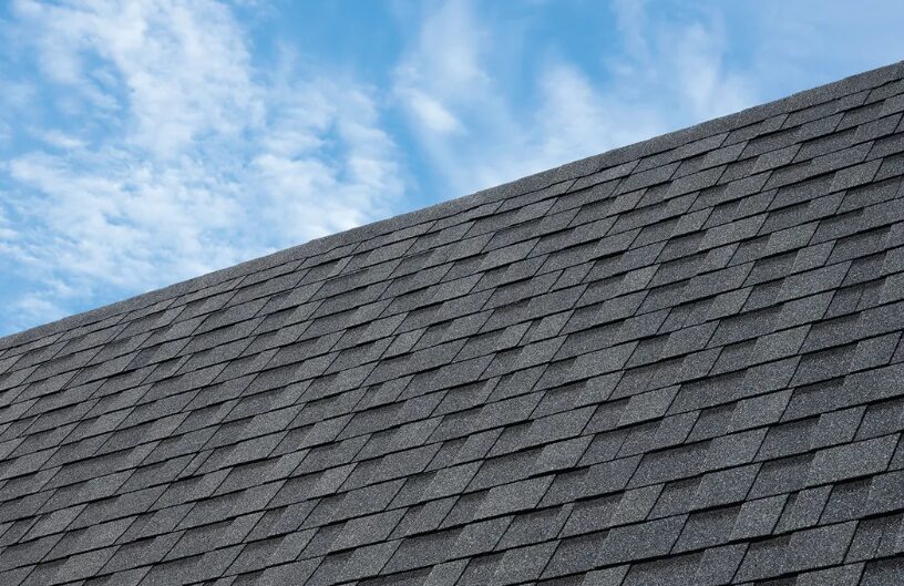 Diagonal view of a textured gray asphalt shingle roof against a blue sky with wispy clouds.