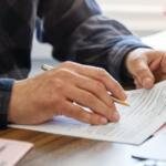 Close-up of hands signing or filling out a form with a gold pen on a wooden desk, paperwork spread out and a small plant in the background