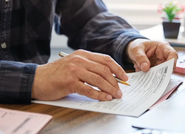 Close-up of hands signing or filling out a form with a gold pen on a wooden desk, paperwork spread out and a small plant in the background