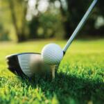 Close-up of a golf ball teed up on a small wooden tee beside a driver on a grassy golf course, sunny day.