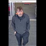 Surveillance image of a man in a dark hoodie and black joggers walking toward the camera at a store entrance, hands in pockets; timestamp shows 05/02/2026 12:04:13 PM.