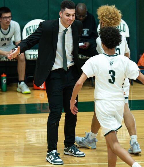 Basketball coach in a black suit and green tie gesturing to a player in a white jersey on a gym floor during a game.
