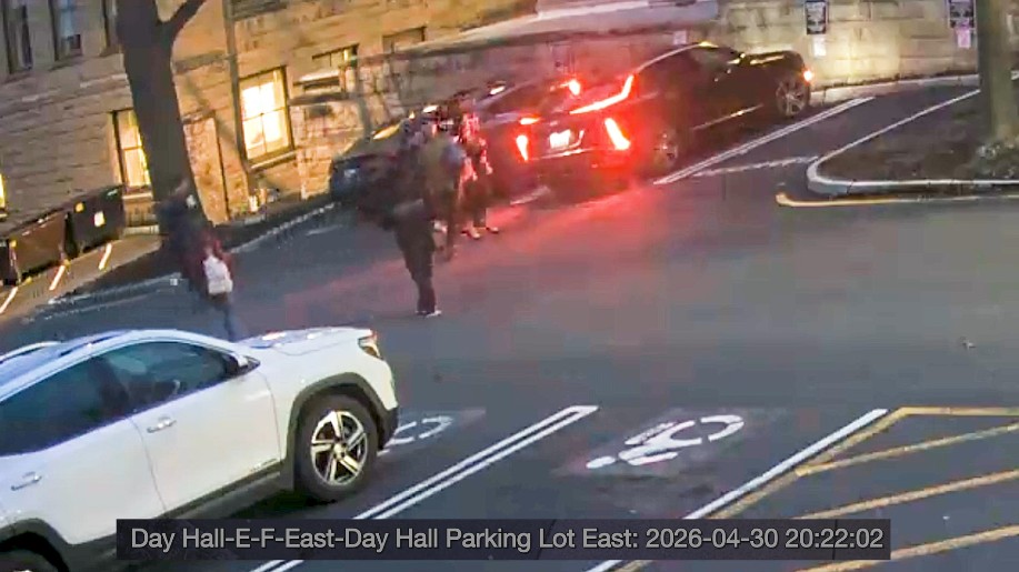 Nighttime parking lot scene with a white SUV in the foreground and a group of people near a car with red brake lights by a stone building.