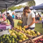 Couple browsing fresh fruit at an outdoor farmers market under tents on a sunny day.