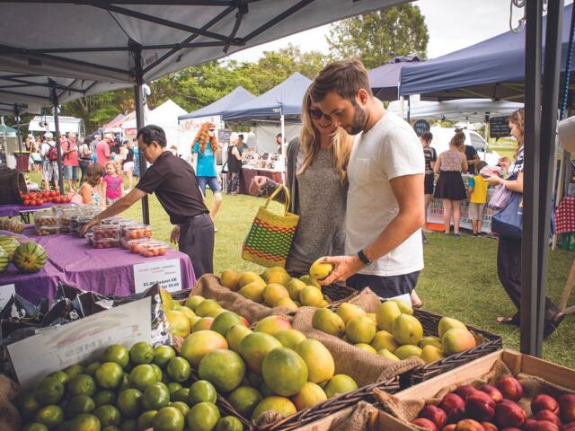 Couple browsing fresh fruit at an outdoor farmers market under tents on a sunny day.