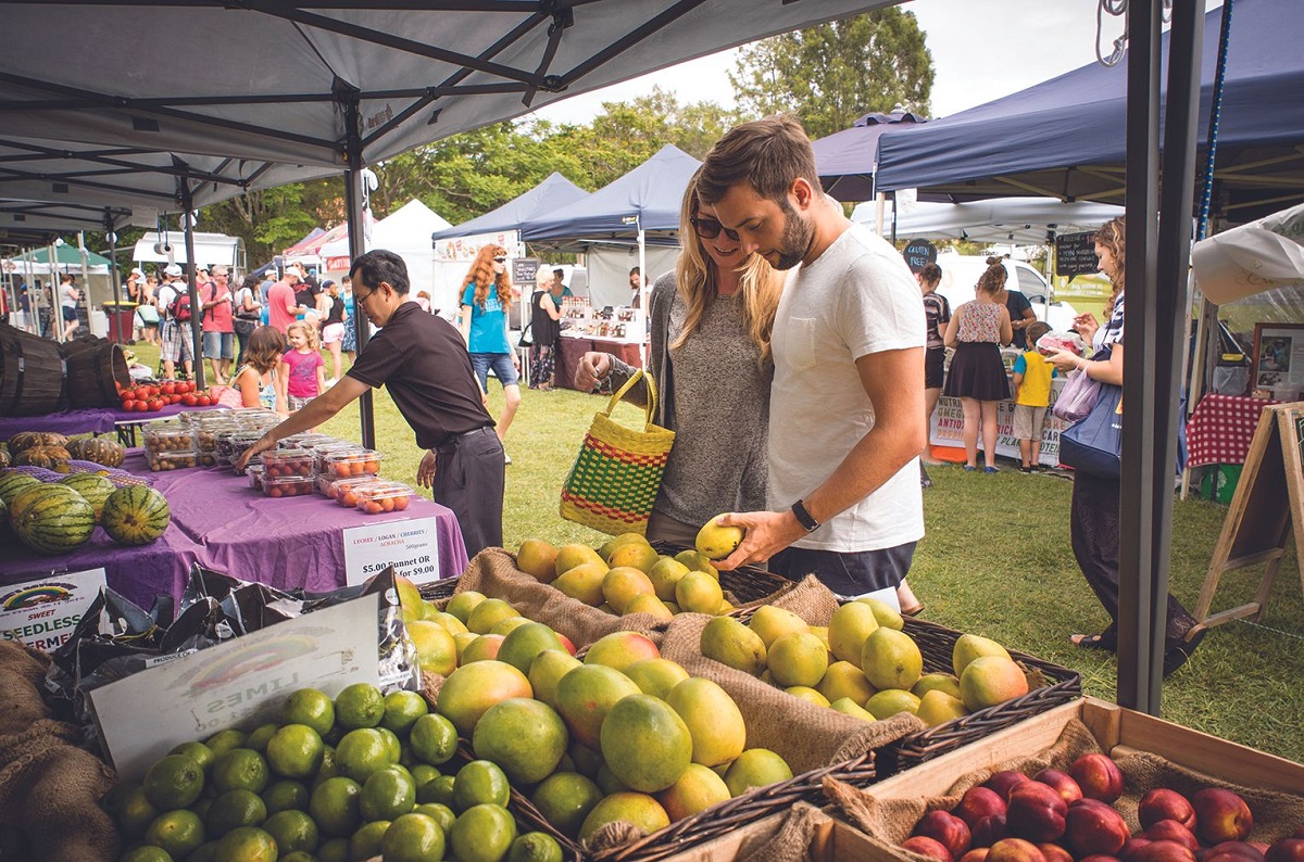 Couple browsing fresh fruit at an outdoor farmers market under tents on a sunny day.