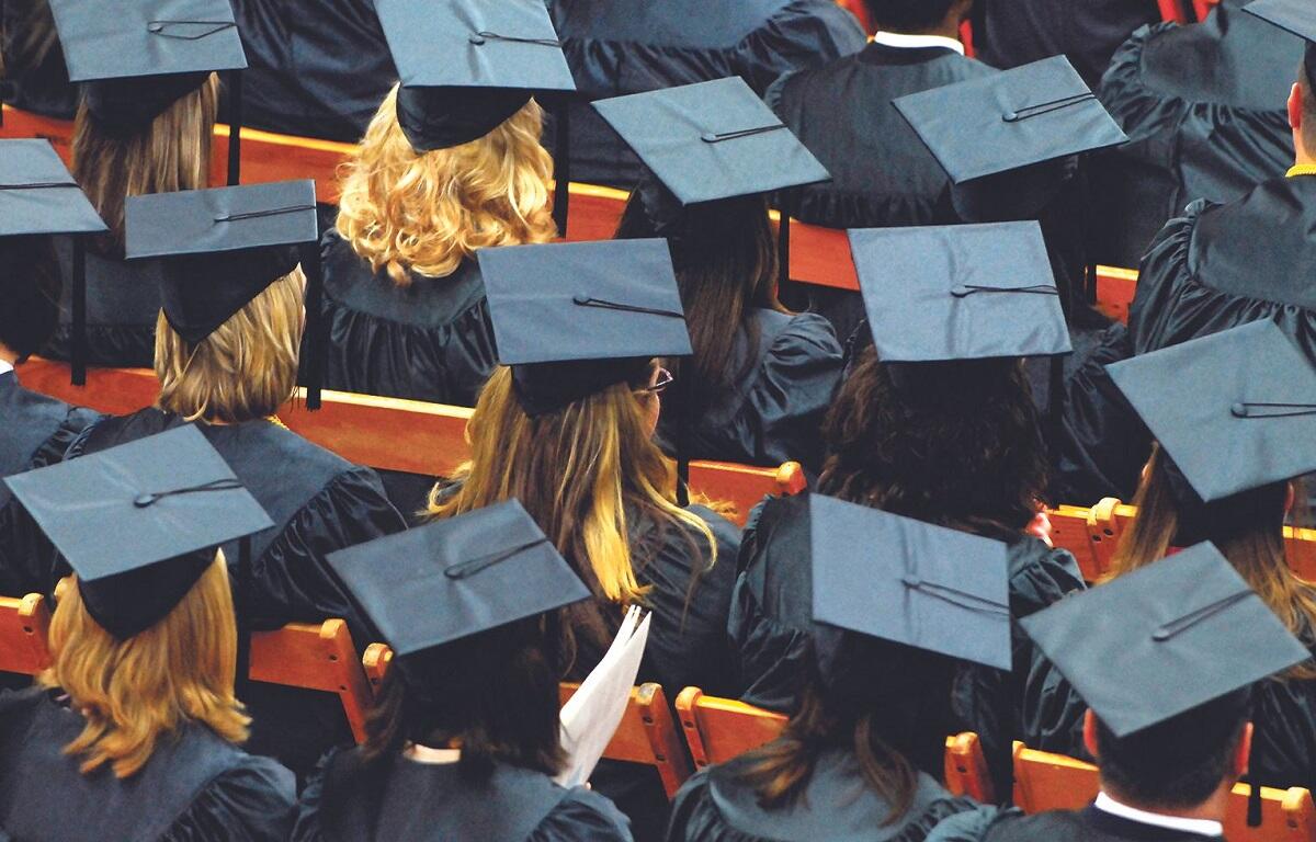 Row of graduates in blue mortarboard caps and gowns seated, viewed from behind at a ceremony.