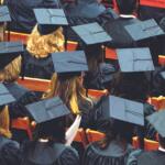 Row of graduates in blue mortarboard caps and gowns seated, viewed from behind at a ceremony.