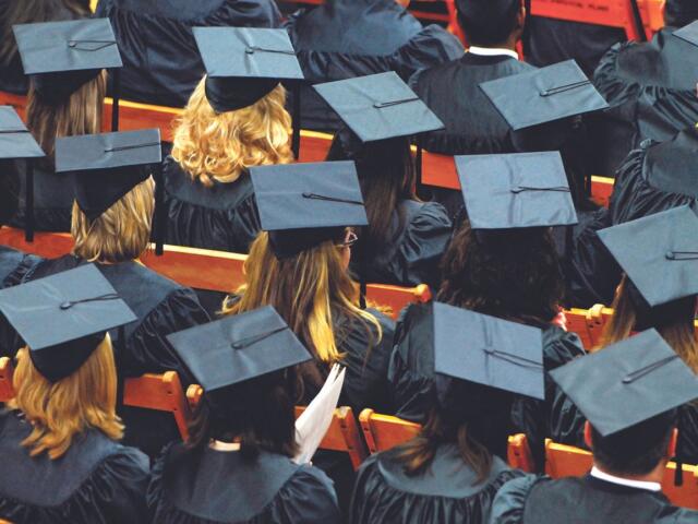 Row of graduates in blue mortarboard caps and gowns seated, viewed from behind at a ceremony.