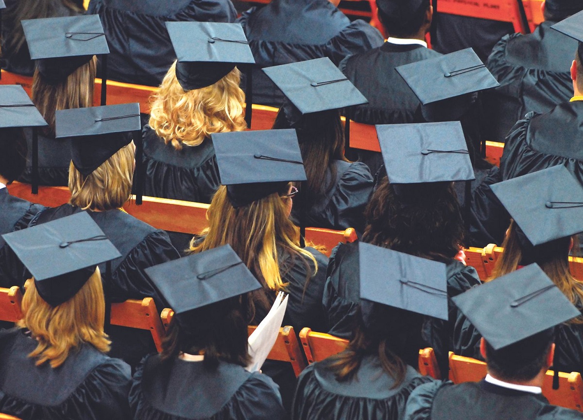 Row of graduates in blue mortarboard caps and gowns seated, viewed from behind at a ceremony.