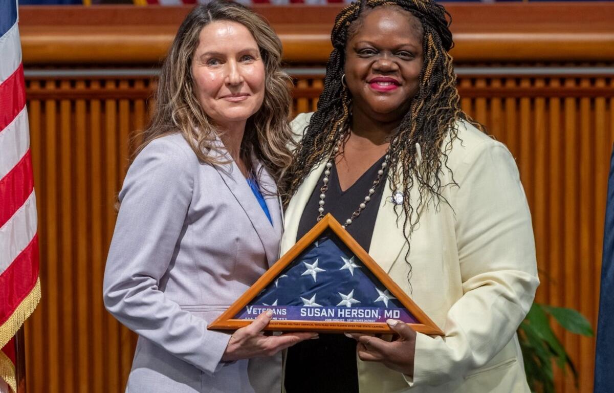 Two women in suits pose on stage, holding a triangular folded American flag plaque that reads 'Veterans Susan Herson, MD'.