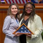 Two women in suits pose on stage, holding a triangular folded American flag plaque that reads 'Veterans Susan Herson, MD'.