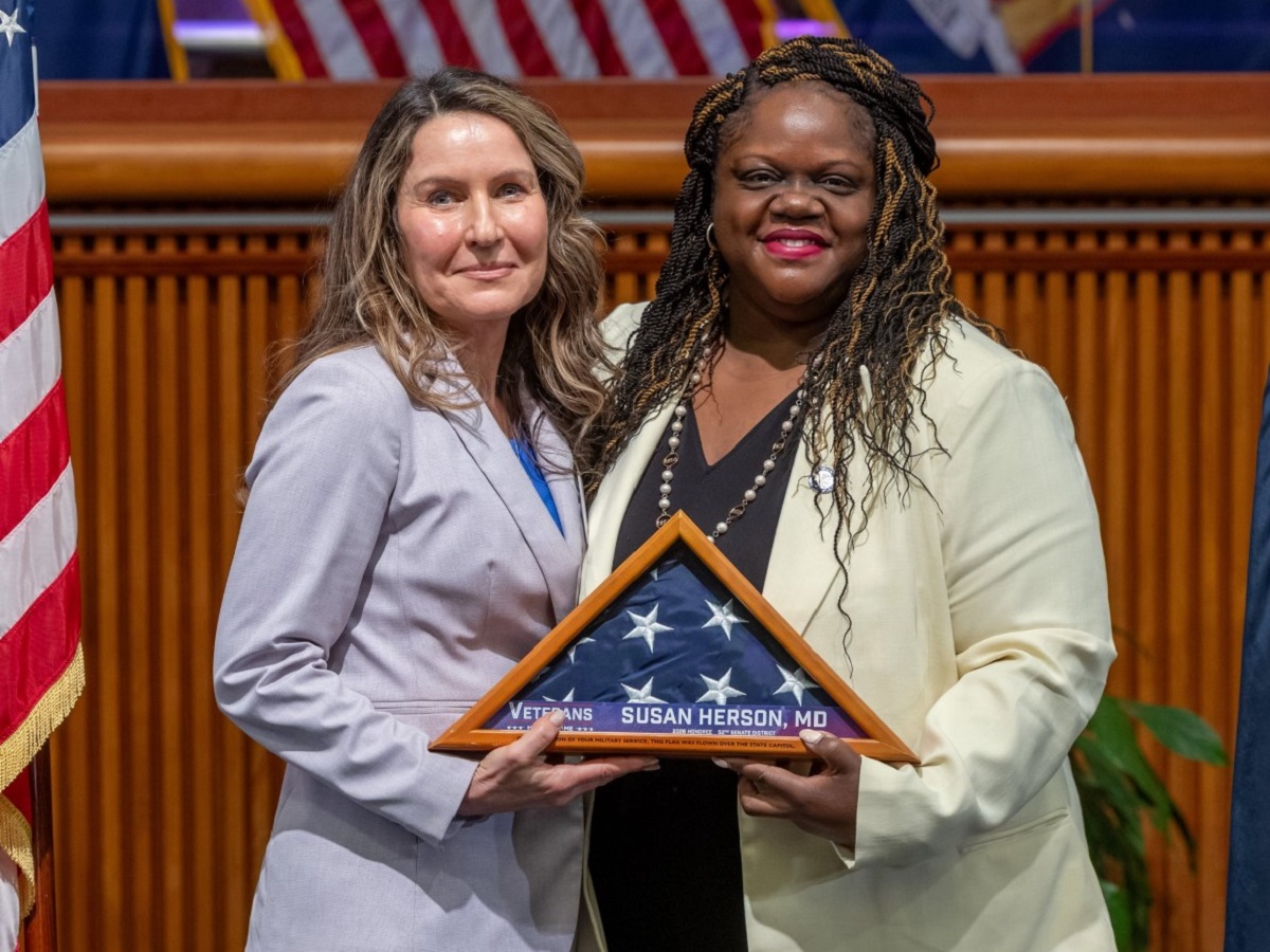 Two women in suits pose on stage, holding a triangular folded American flag plaque that reads 'Veterans Susan Herson, MD'.