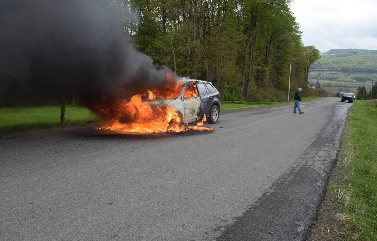 SUV on fire in the middle of a rural road with thick black smoke; flames engulf the front and cabin as a man in a cap walks away toward the right and a second car approaches in the distance.