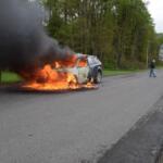 SUV on fire in the middle of a rural road with thick black smoke; flames engulf the front and cabin as a man in a cap walks away toward the right and a second car approaches in the distance.