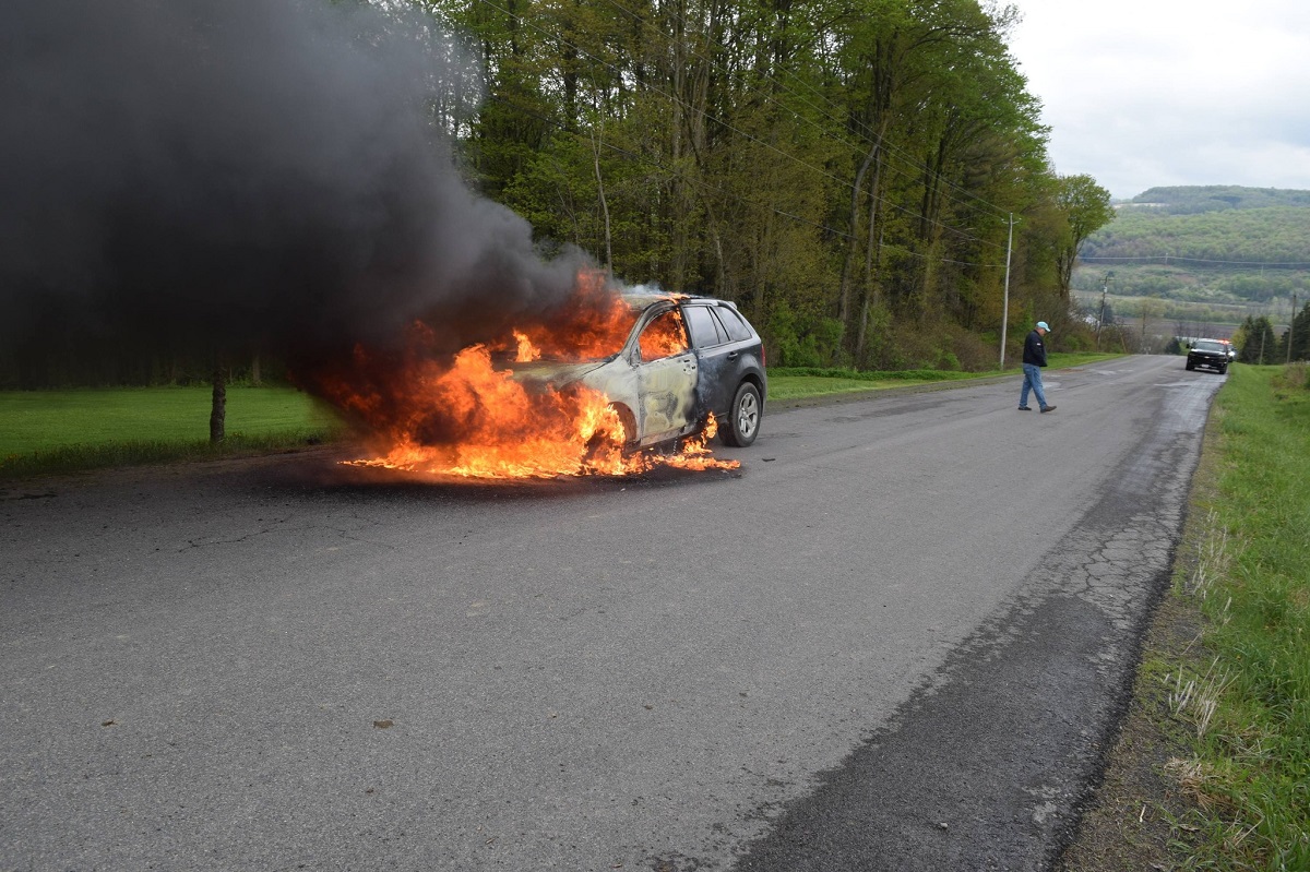 SUV on fire in the middle of a rural road with thick black smoke; flames engulf the front and cabin as a man in a cap walks away toward the right and a second car approaches in the distance.
