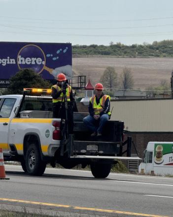 Two roadside construction workers in reflective vests and red helmets stand and sit on a flatbed truck by a roadway, with a 'begin here' billboard in the background.
