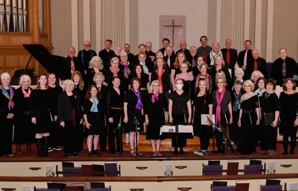 Large choir of men and women in black clothing, many wearing colorful scarves, posing on church choir risers with a cross on the wall behind them and organ pipes to the left.