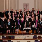Large choir of men and women in black clothing, many wearing colorful scarves, posing on church choir risers with a cross on the wall behind them and organ pipes to the left.