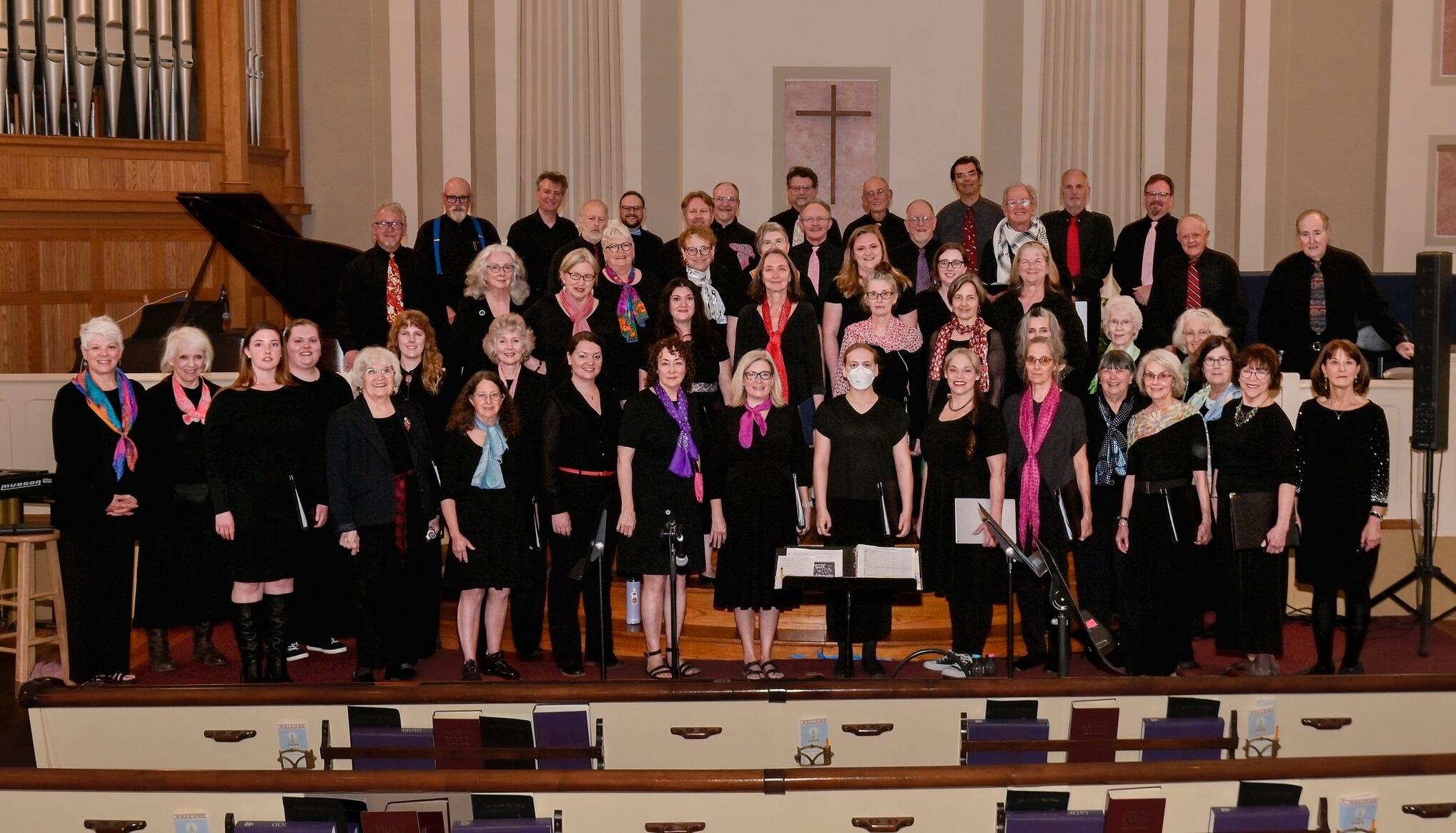 Large choir of men and women in black clothing, many wearing colorful scarves, posing on church choir risers with a cross on the wall behind them and organ pipes to the left.