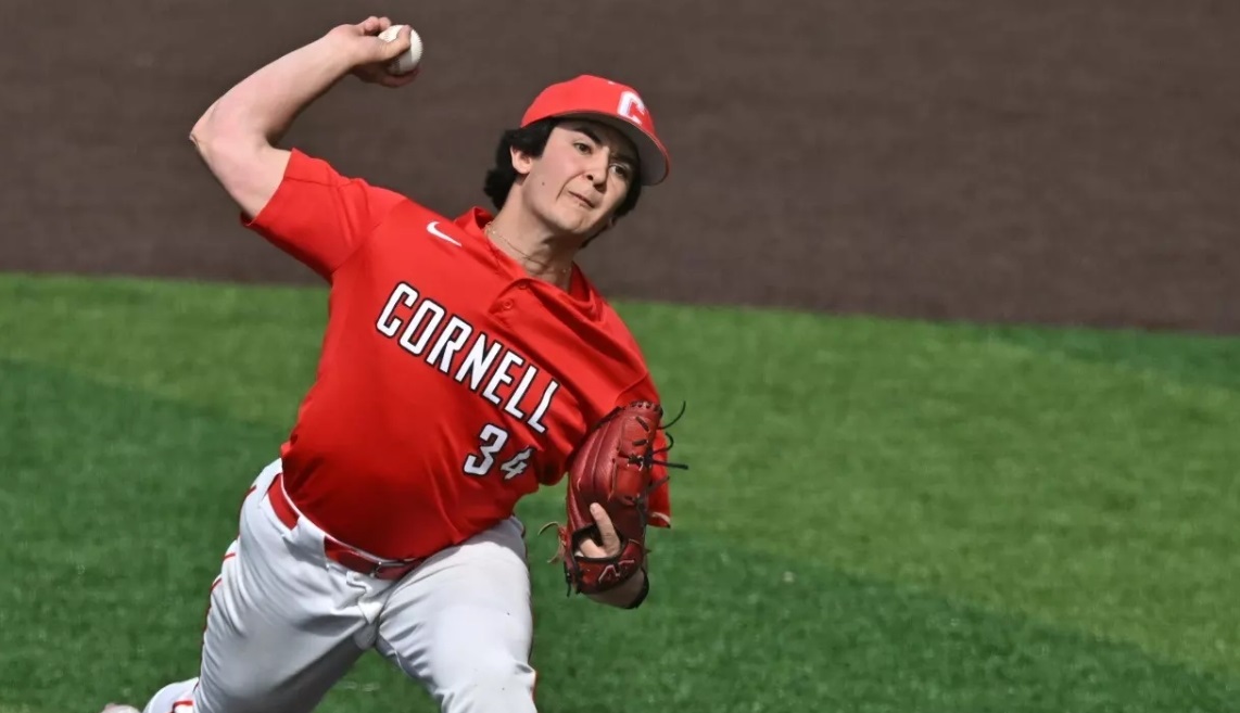 Female baseball pitcher in a Cornell red uniform (number 34) throws a pitch on the mound, glove on left hand and ball raised overhead.