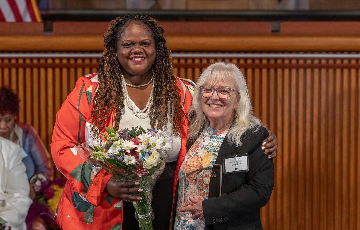 Two smiling women pose together at a formal event; the woman on the left holds a bouquet of flowers.