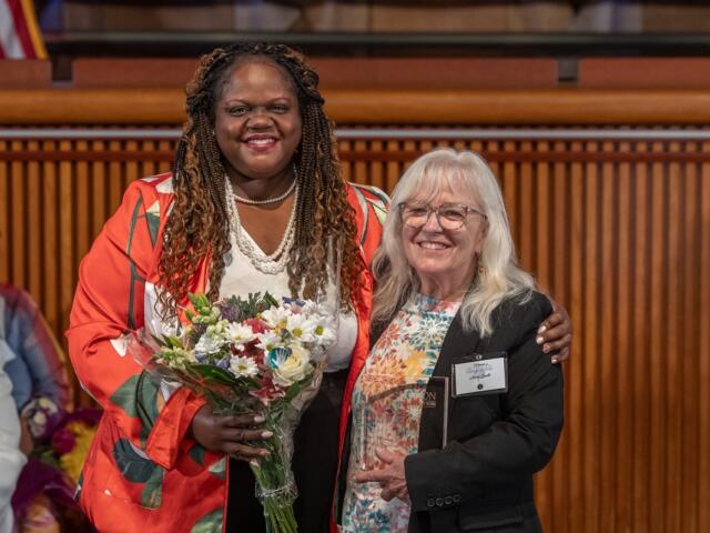 Two smiling women pose together at a formal event; the woman on the left holds a bouquet of flowers.