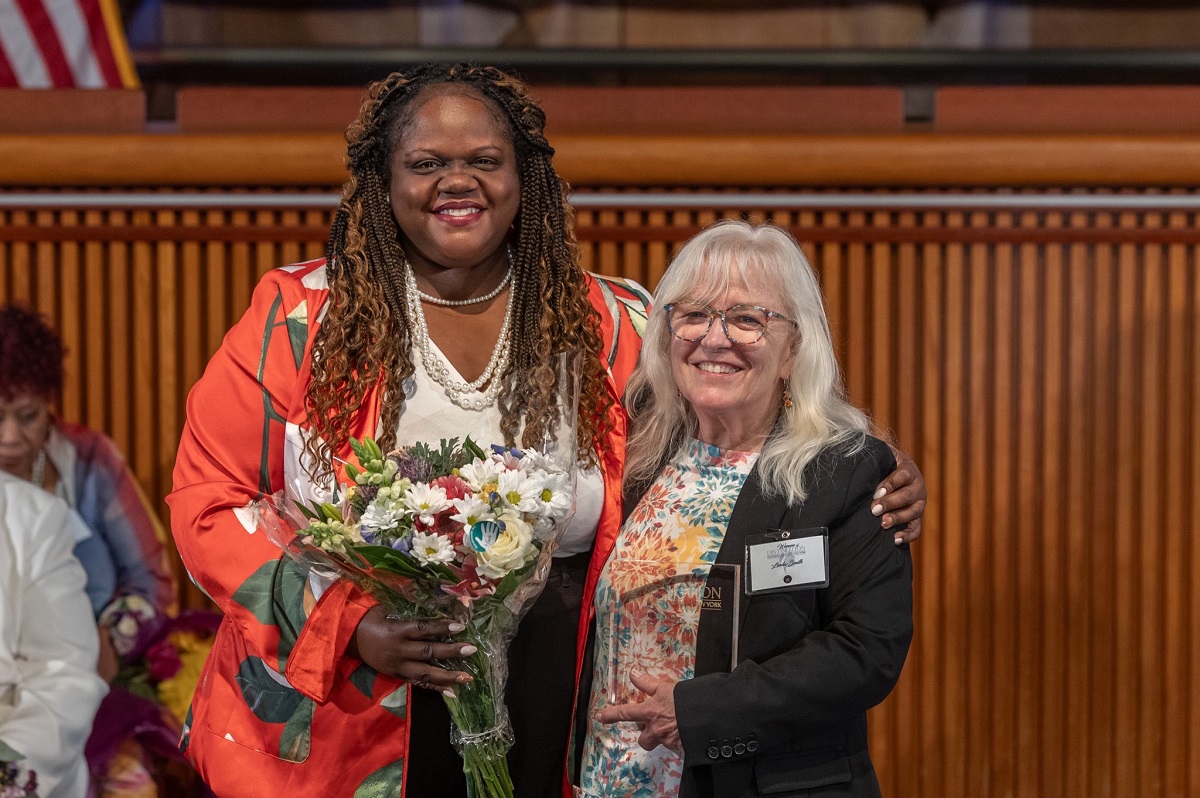 Two smiling women pose together at a formal event; the woman on the left holds a bouquet of flowers.
