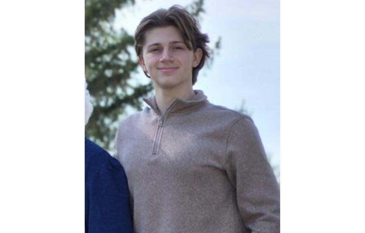 Young person standing outdoors in daylight, wearing a light brown zip-neck sweater and smiling at the camera.