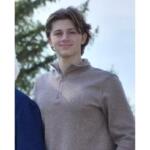 Young person standing outdoors in daylight, wearing a light brown zip-neck sweater and smiling at the camera.