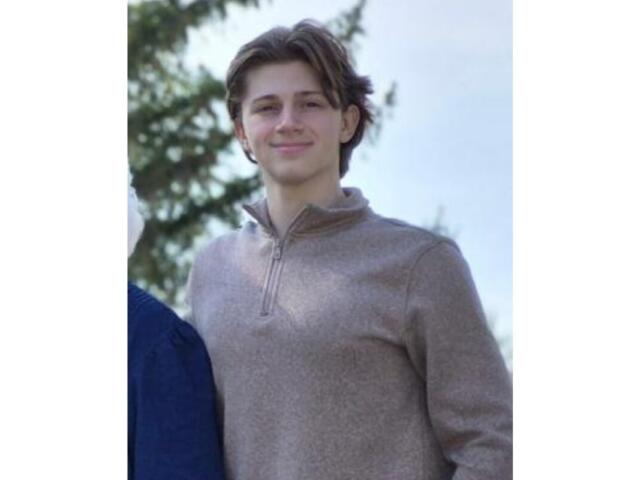 Young person standing outdoors in daylight, wearing a light brown zip-neck sweater and smiling at the camera.