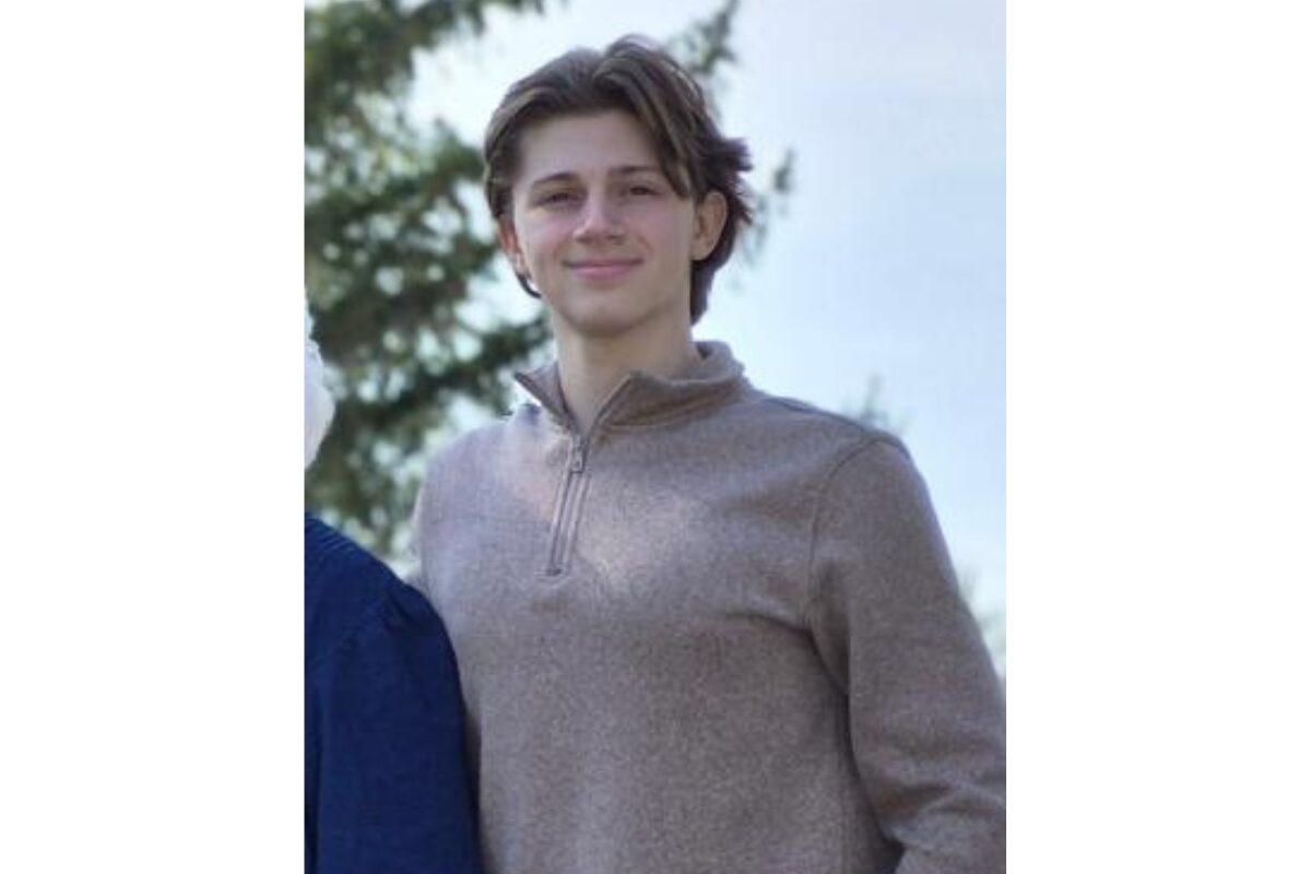 Young person standing outdoors in daylight, wearing a light brown zip-neck sweater and smiling at the camera.