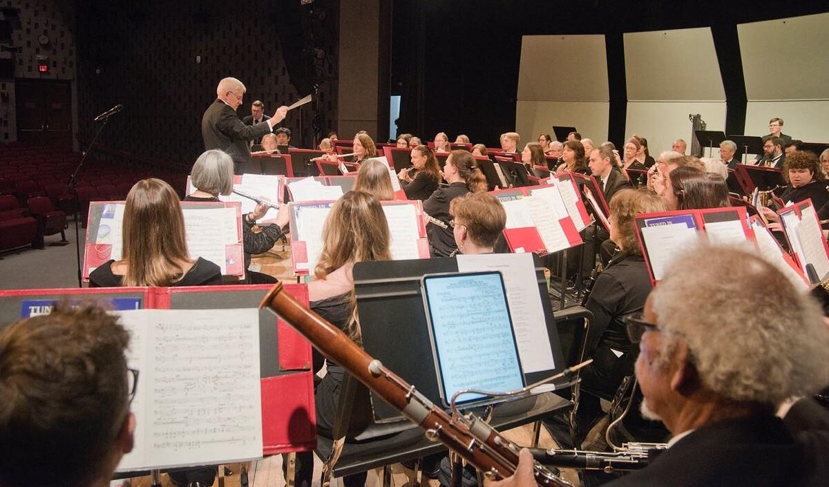 Conductor leads a large orchestra on a stage, musicians reading sheet music and playing instruments in a concert hall.