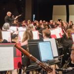 Conductor leads a large orchestra on a stage, musicians reading sheet music and playing instruments in a concert hall.