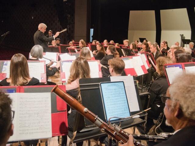 Conductor leads a large orchestra on a stage, musicians reading sheet music and playing instruments in a concert hall.
