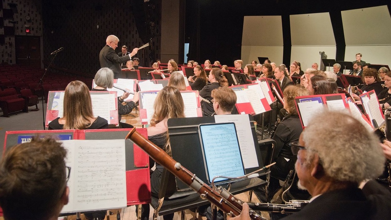 Conductor leads a large orchestra on a stage, musicians reading sheet music and playing instruments in a concert hall.