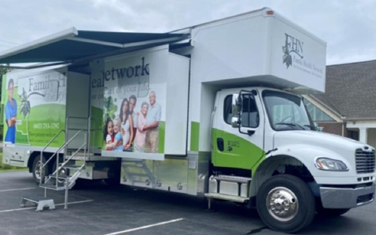 White and green mobile health clinic truck with its side banners showing a family and outreach graphics, awning extended and stairs deployed in a parking lot.
