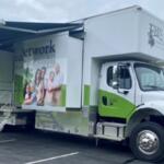 White and green mobile health clinic truck with its side banners showing a family and outreach graphics, awning extended and stairs deployed in a parking lot.