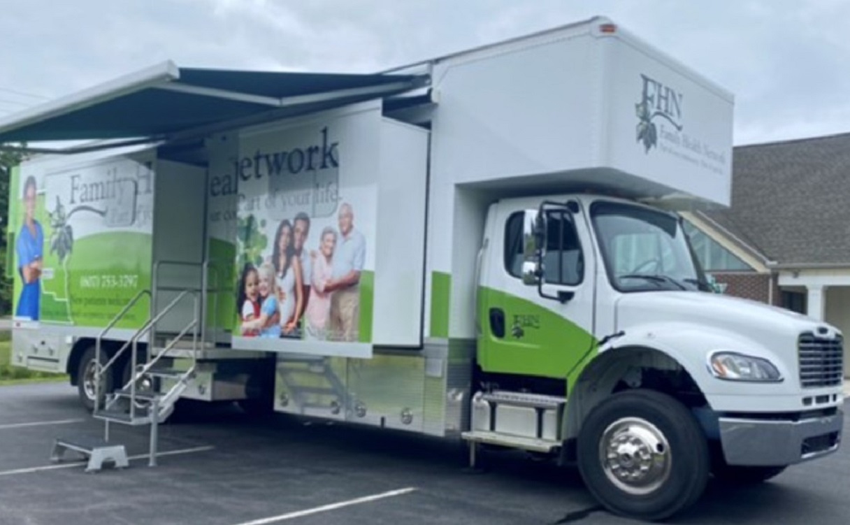 White and green mobile health clinic truck with its side banners showing a family and outreach graphics, awning extended and stairs deployed in a parking lot.