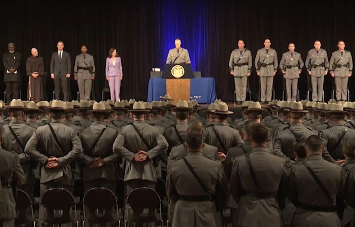 Stage ceremony with a speaker at a podium, officials to the sides, and rows of uniformed soldiers standing with hands behind their backs facing an audience in a large hall.