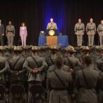 Stage ceremony with a speaker at a podium, officials to the sides, and rows of uniformed soldiers standing with hands behind their backs facing an audience in a large hall.