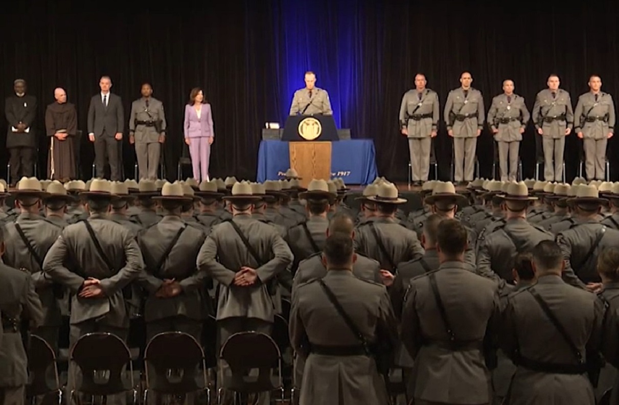 Stage ceremony with a speaker at a podium, officials to the sides, and rows of uniformed soldiers standing with hands behind their backs facing an audience in a large hall.