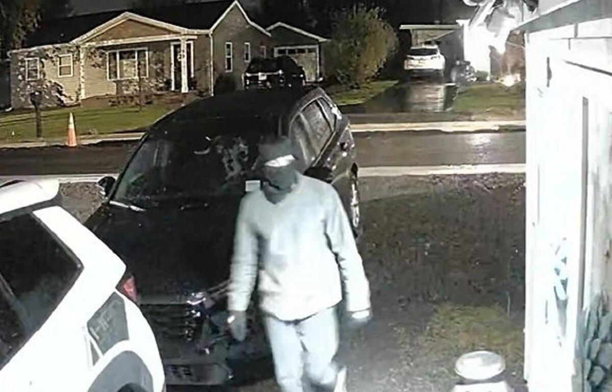 Person in a gray hoodie walks toward a dark SUV parked in a residential driveway at night under wet pavement lighting