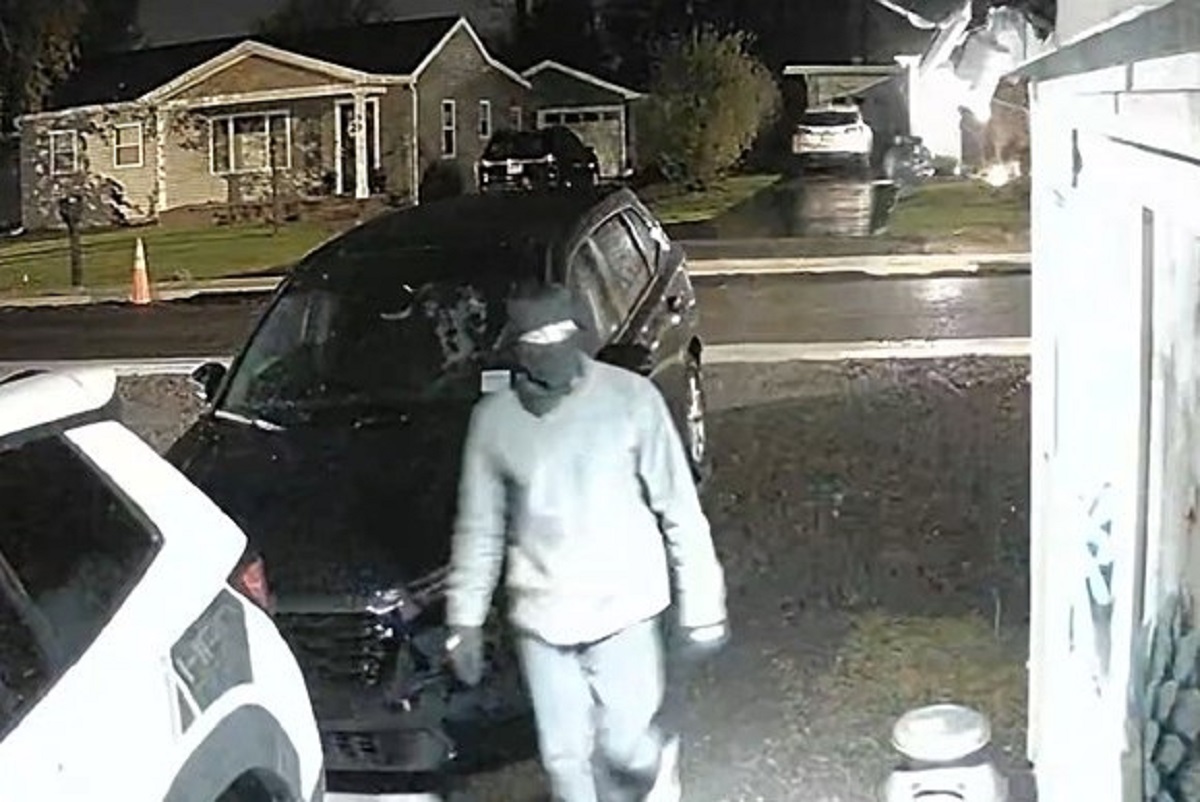 Person in a gray hoodie walks toward a dark SUV parked in a residential driveway at night under wet pavement lighting